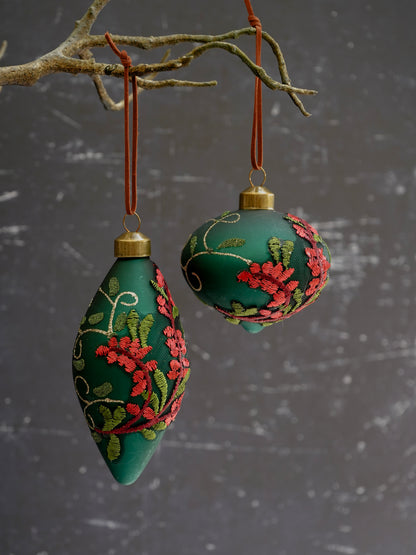 Two green embroidered ornaments hanging from branches against a gray background