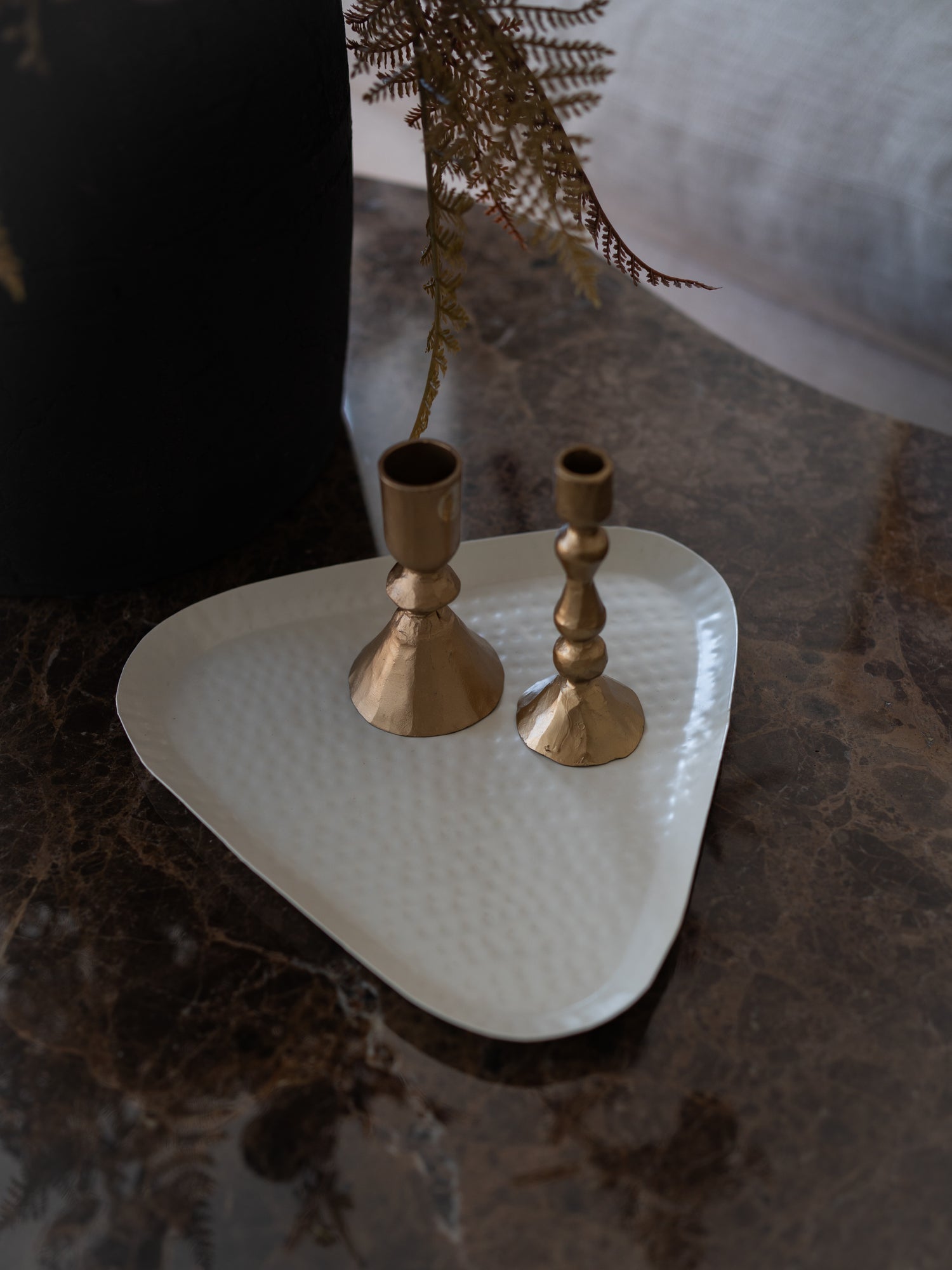 Two brass candlesticks of different heights displayed on a white hammered metal tray, set on a dark marble surface.