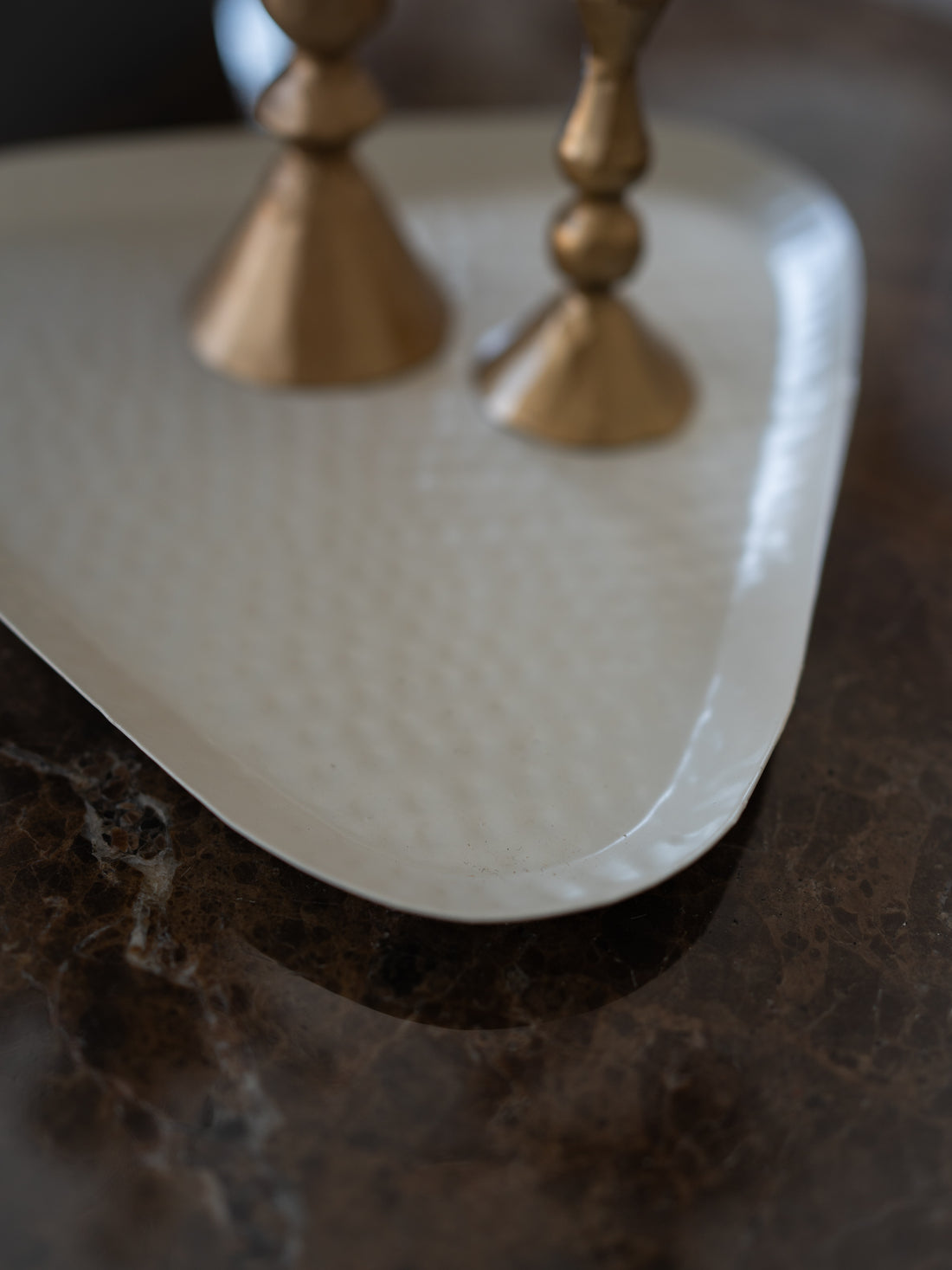 A close-up of a white hammered metal serving tray with curved edges, resting on a dark brown marble surface with two gold-toned candlesticks partially visible in the background.