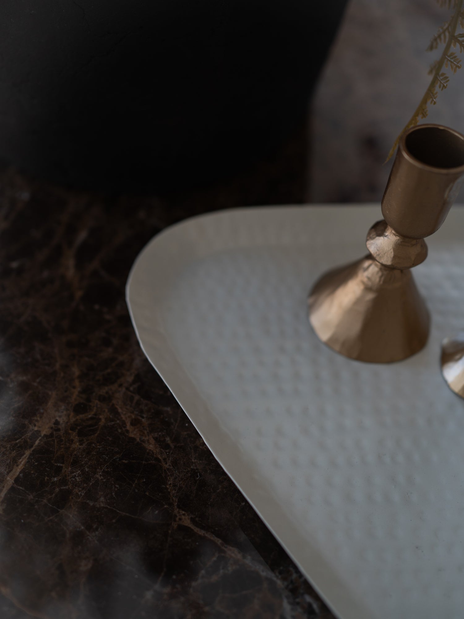 A close-up of a hammered white metal tray on a dark marble surface, featuring a brass candlestick holder and part of another metallic object.