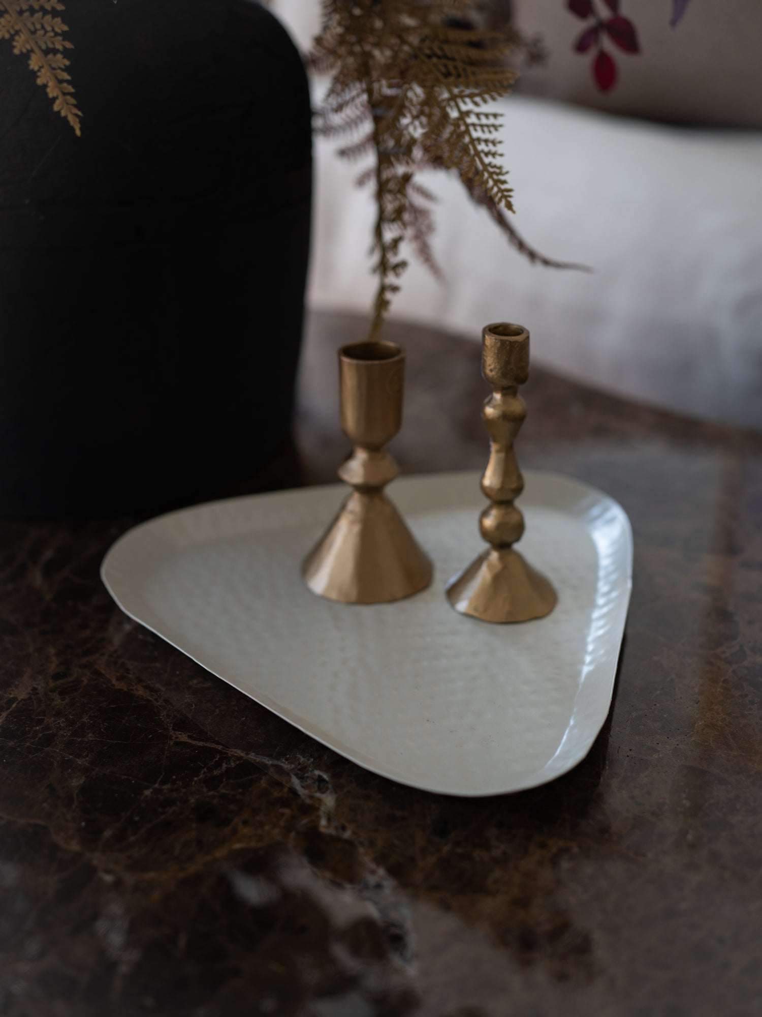 Two brass candlesticks of different heights standing on a white triangular tray, placed on a dark marble surface with a blurred black vase and dried fern in the background.