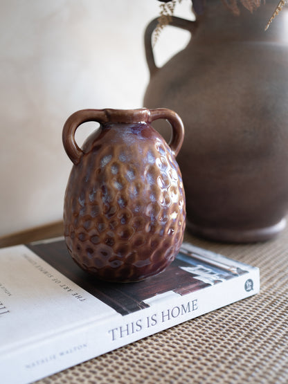 Small mulberry textured vase on a book with a larger brown vase in the background.