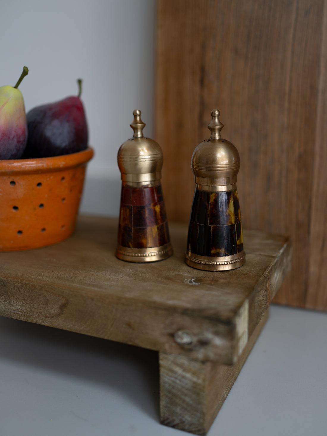 Two decorative brass and tortoiseshell shakers on a wooden surface with a basket of fruits in the background.