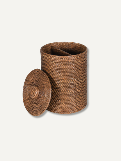 A natural brown woven rattan storage bin with a matching round lid leaning against it, set against a solid off-white background.
