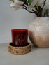 Red candle with branding on a woven coaster next to a textured vase and flowers.