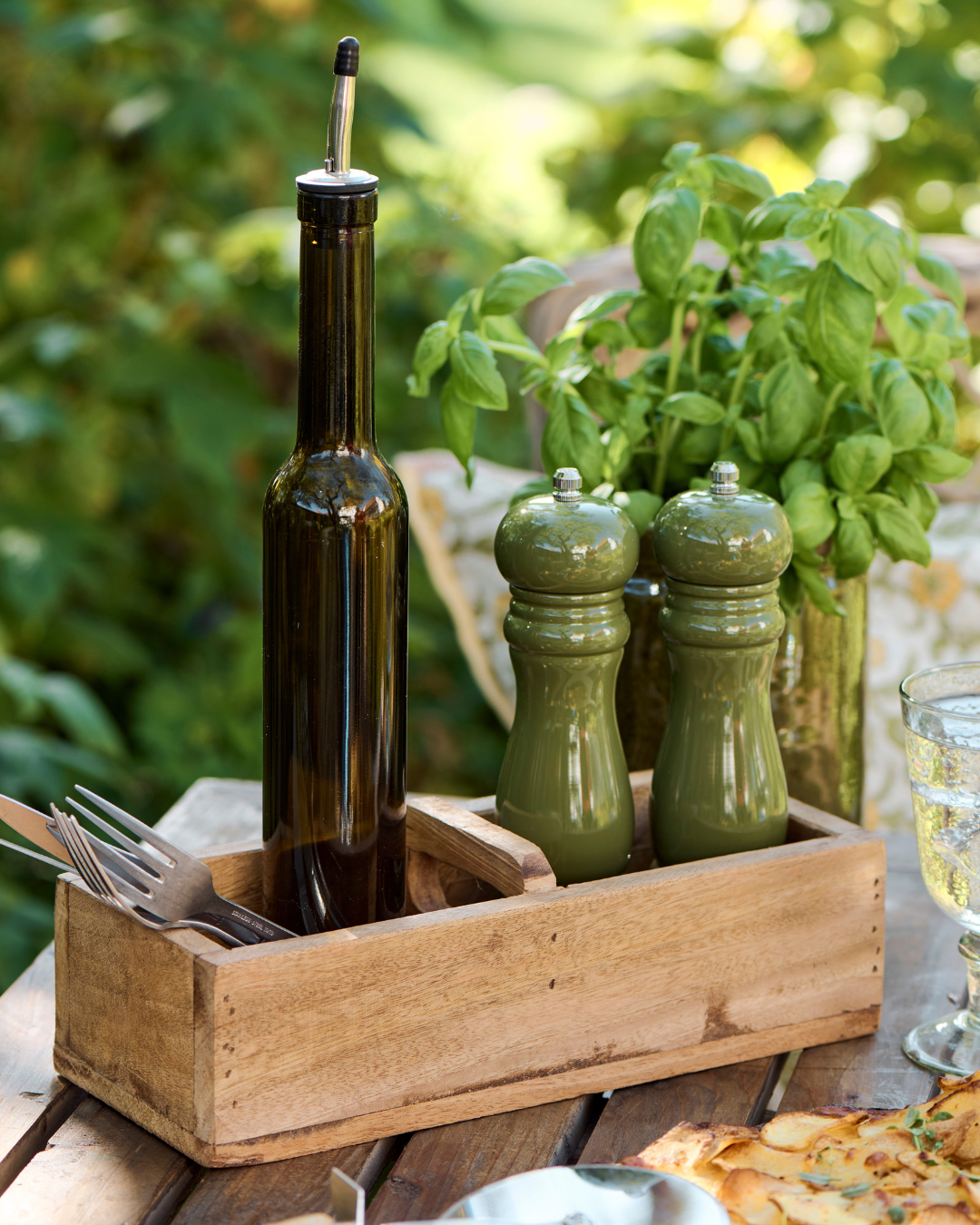 Olive oil bottle, salt and pepper mills in a wooden box on a table with greenery in the background