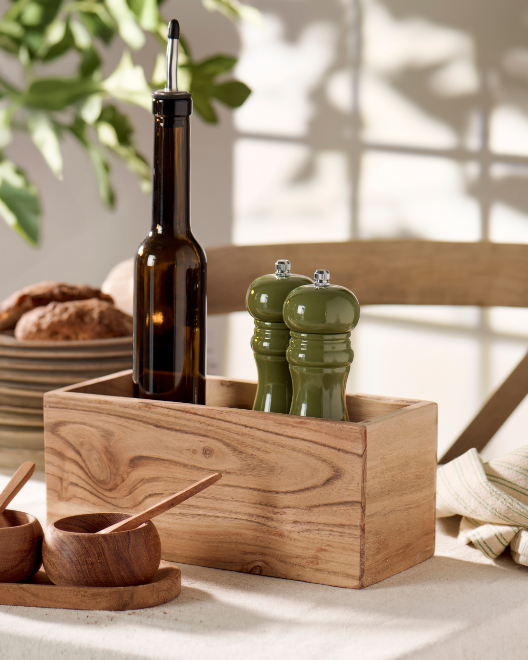 Wooden box with olive oil bottle, salt and pepper mills, and wooden spoons on a light surface.