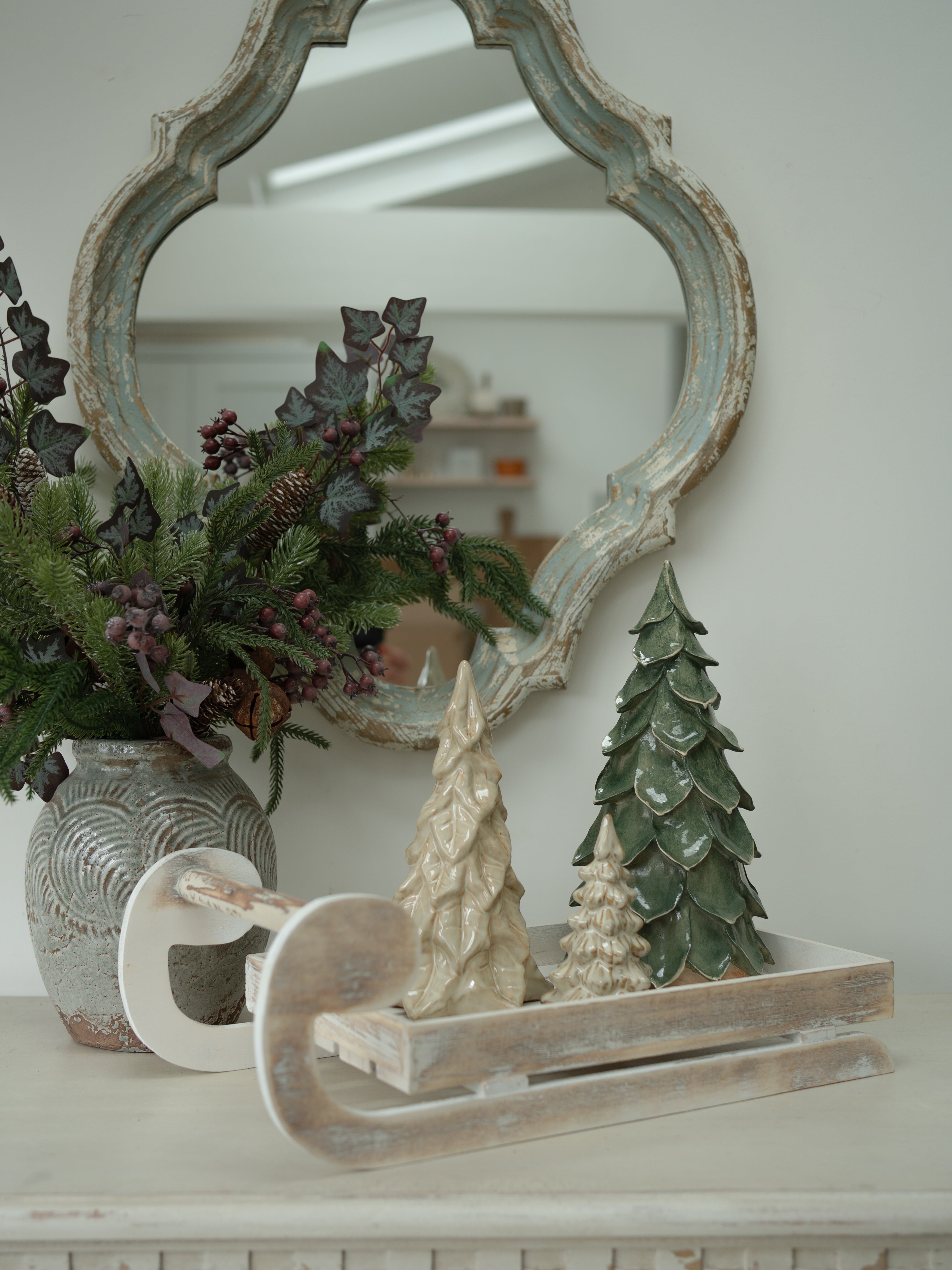 A rustic holiday tabletop arrangement featuring a distressed white wooden sled with decorative Christmas tree figurines, a textured gray ceramic vase filled with green pine garlands and berries, and a large ornate distressed mirror in the background.