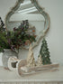 A rustic holiday tabletop arrangement featuring a distressed white wooden sled with decorative Christmas tree figurines, a textured gray ceramic vase filled with green pine garlands and berries, and a large ornate distressed mirror in the background.