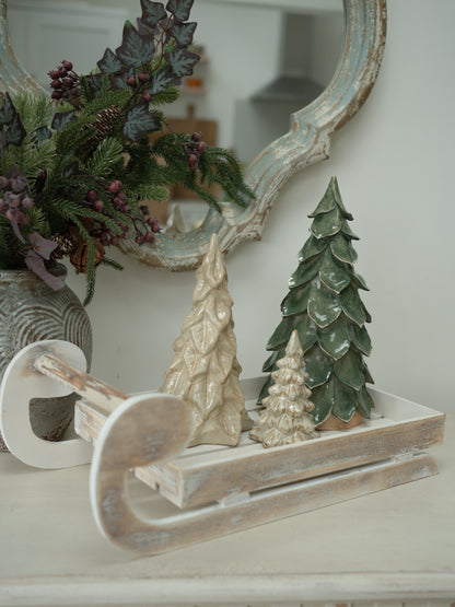 A christmas decorative set featuring a distressed white wooden sled with three textured Christmas tree figurines in cream and green, styled on a mantelpiece next to a mirror and a floral arrangement.