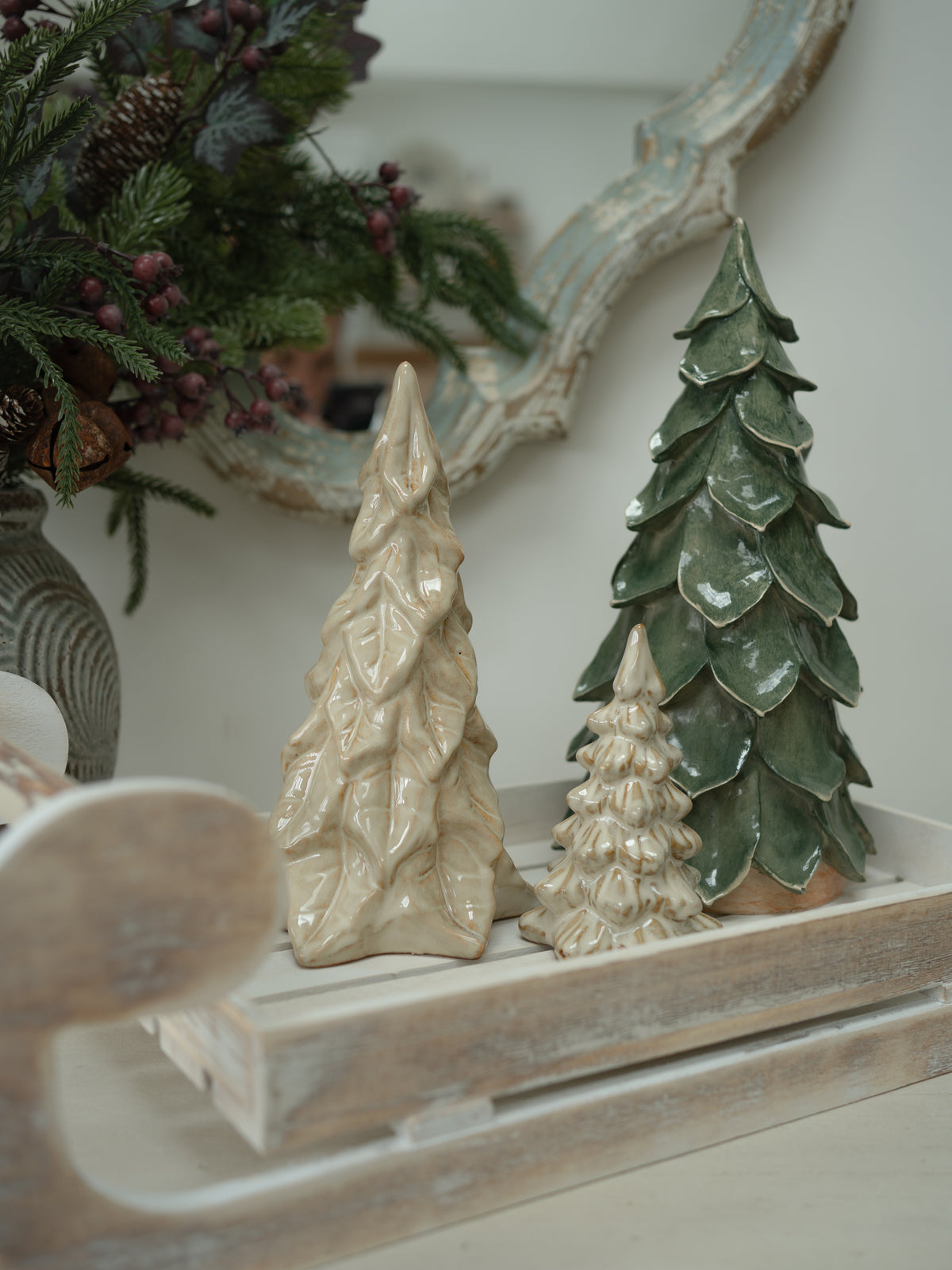Three textured ceramic Christmas tree ornaments in cream and green glazes displayed on a rustic white wooden tray.