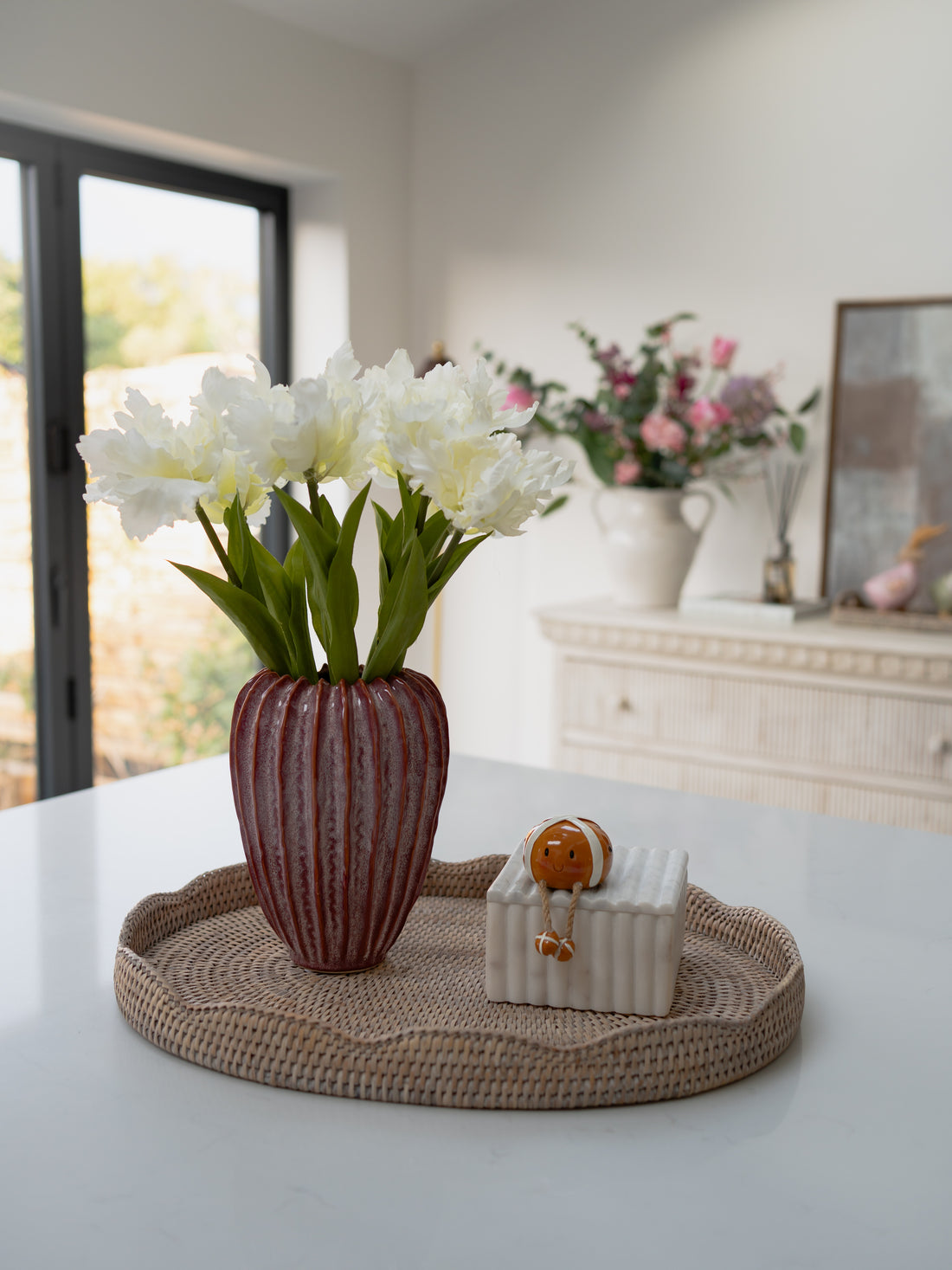 White tulips in a striped vase on a woven tray with a small box, set against a blurred indoor background.