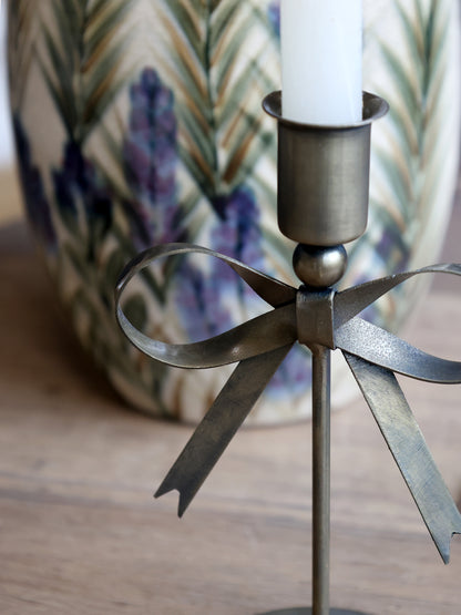 Decorative metal bow candle holder with a candle on a wooden surface, with a blurred floral-patterned vase in the background.