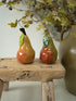 Two glazed ceramic pears with green leaves sit on a rustic wooden stool, with a large vase and dried branches in the background.