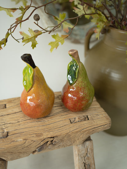 Two glazed ceramic pears with green leaves and dark stems, displayed on a rustic wooden stool with a blurred vase and branches in the background.