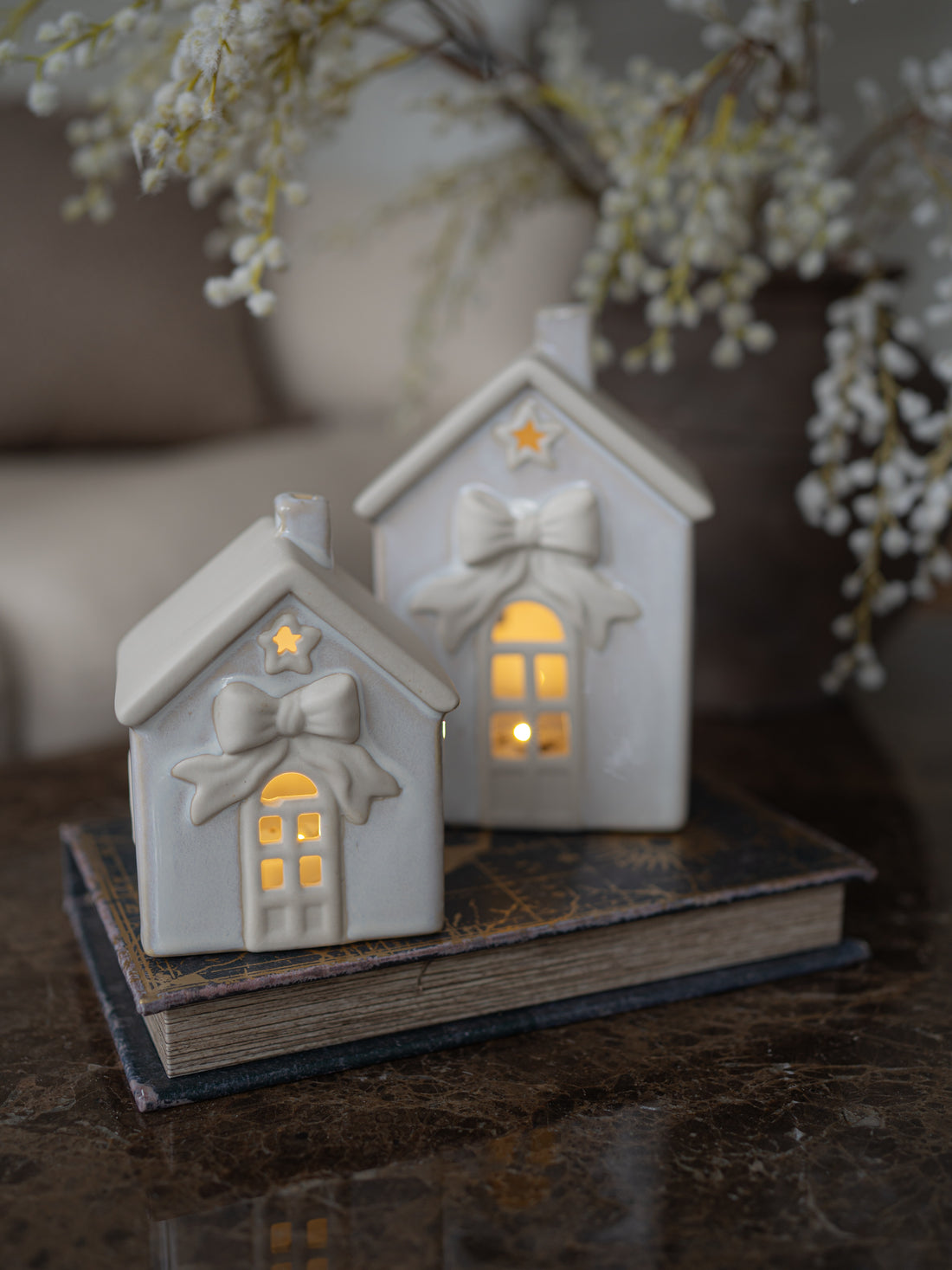 Two small white ceramic house-shaped candle holders with bow and star details, lit from within, displayed on a vintage book on a dark marble surface with blurred white flowers in the background.