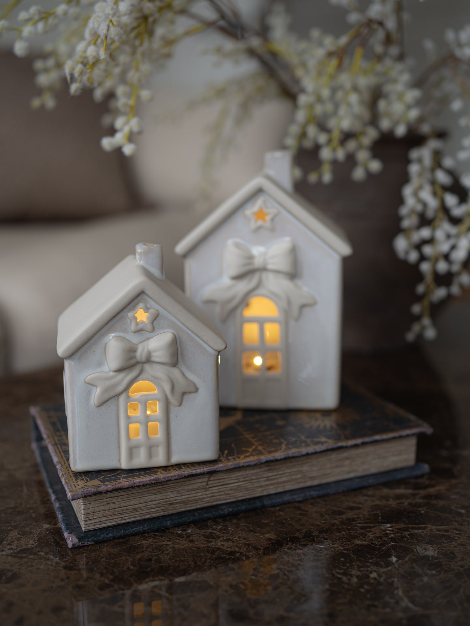 Two small white ceramic house-shaped candle holders with bow and star details, lit from within, displayed on a vintage book on a dark marble surface with blurred white flowers in the background.