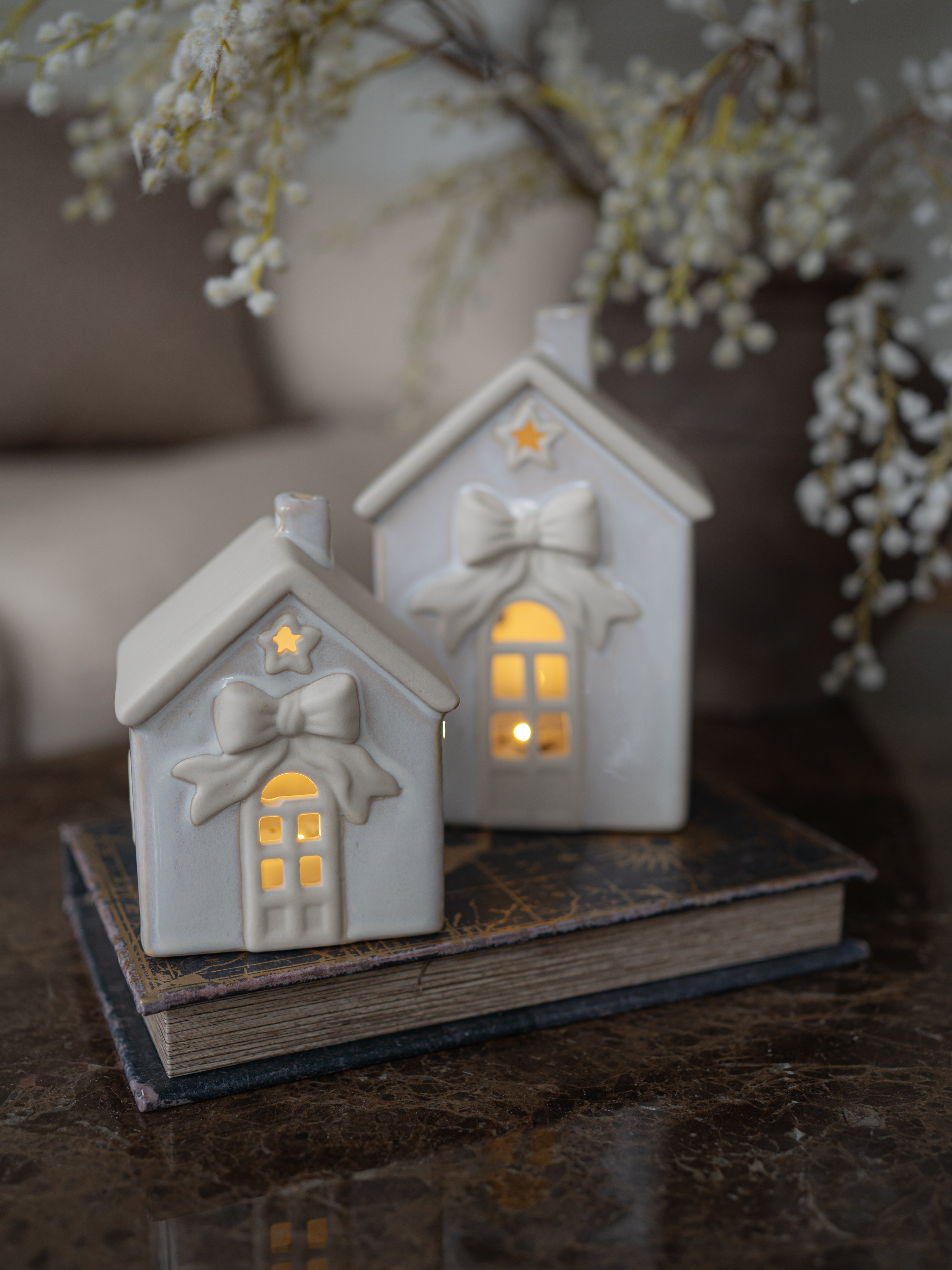 Two small white ceramic house-shaped candle holders with bow and star details, lit from within, displayed on a vintage book on a dark marble surface with blurred white flowers in the background.