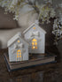 Two small white ceramic house-shaped candle holders with bow and star details, lit from within, displayed on a vintage book on a dark marble surface with blurred white flowers in the background.
