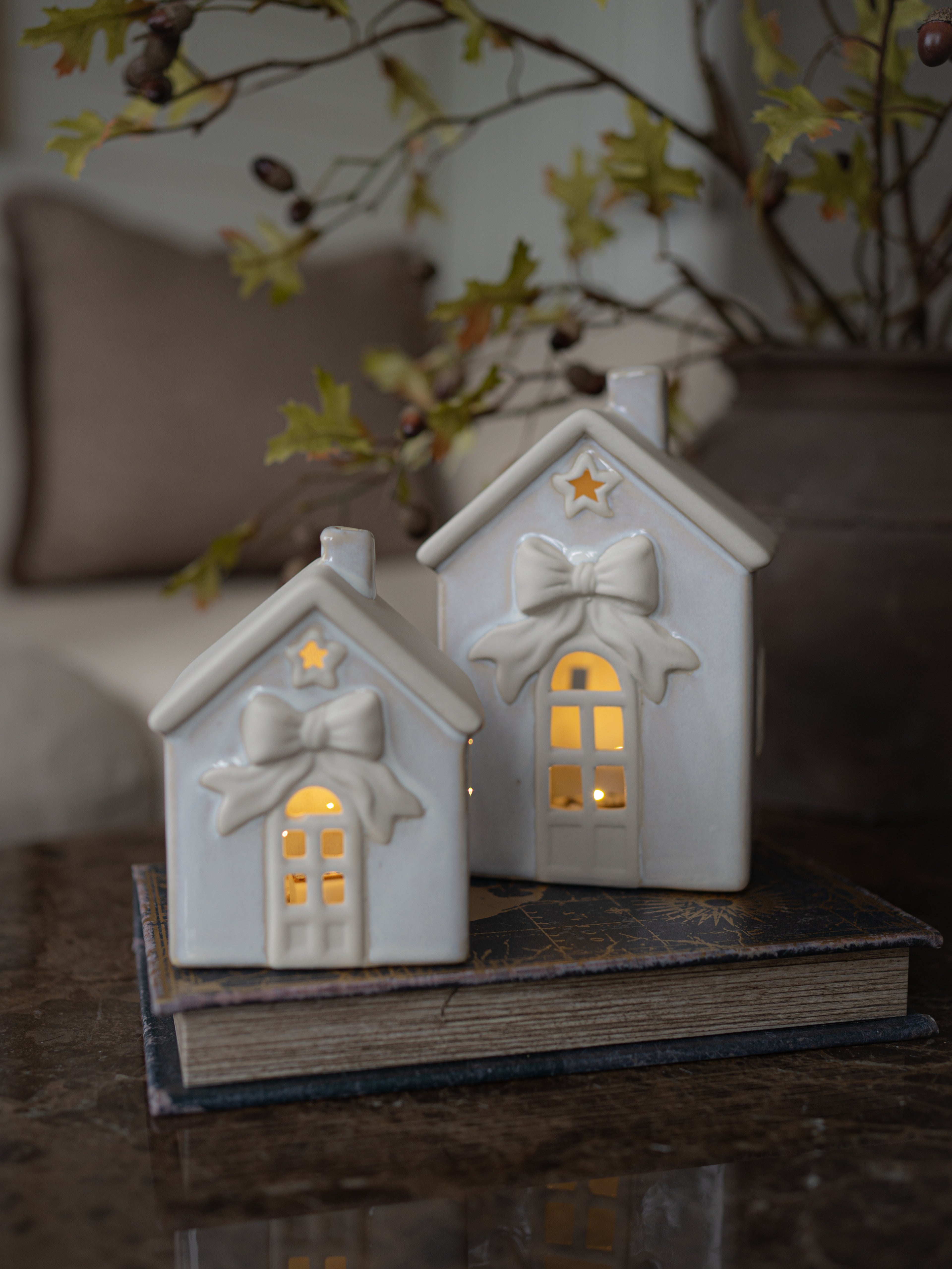 Two small white ceramic house-shaped candle holders with glowing warm lights inside, decorated with bow and star details, displayed on top of a vintage book in a cozy indoor setting.