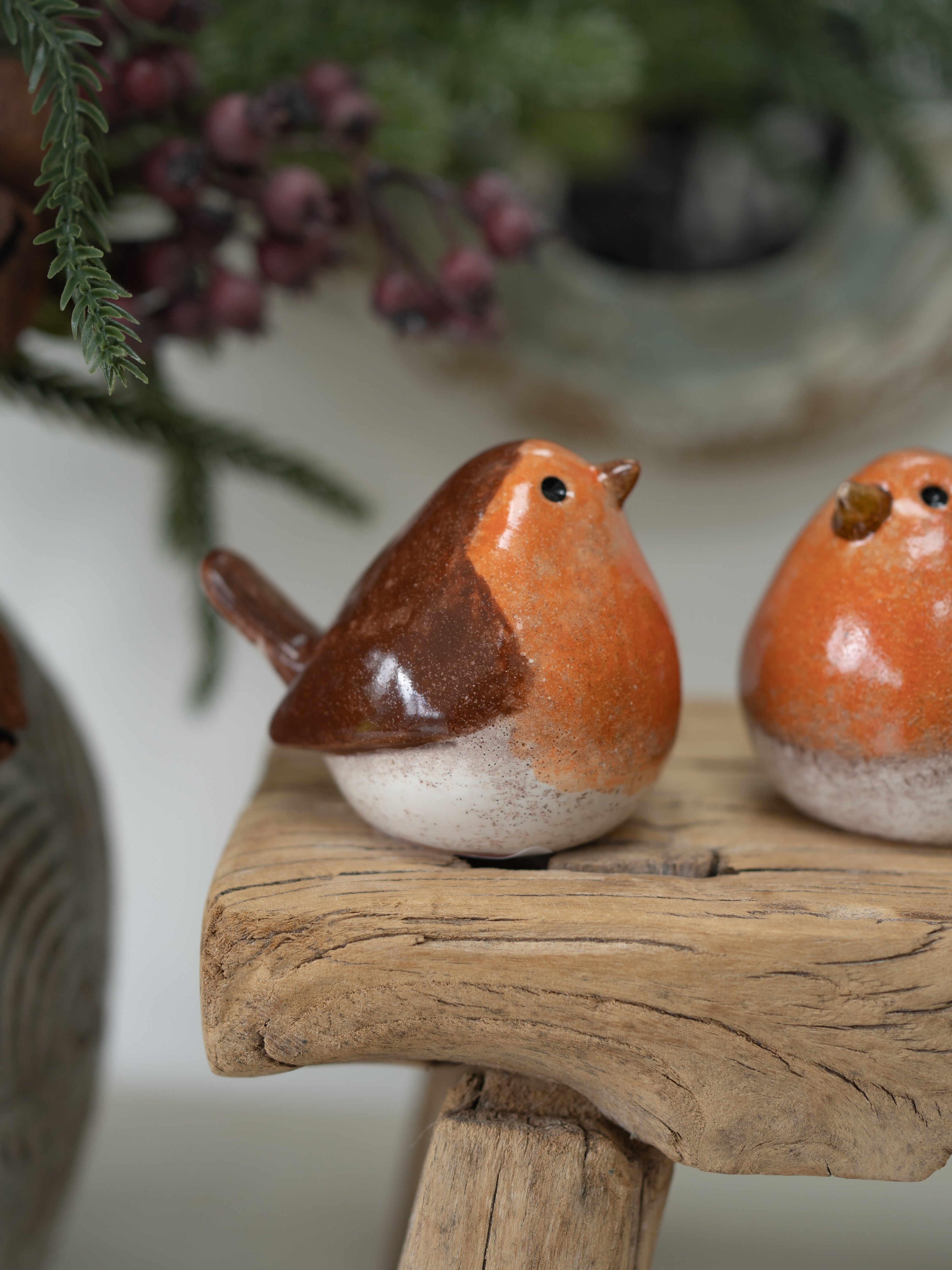 Two small glazed robin bird figurines in orange, brown, and off-white ceramic, displayed on a rustic wooden stand with a softly blurred holiday background.