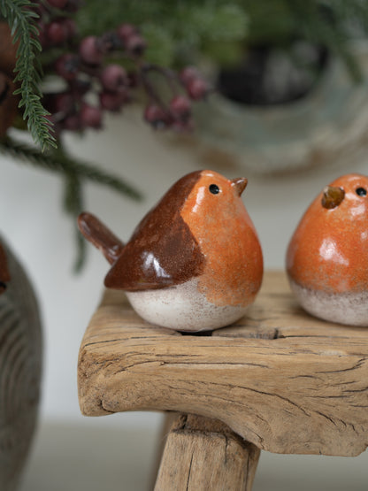 Two small glazed robin bird figurines in orange, brown, and off-white ceramic, displayed on a rustic wooden stand with a softly blurred holiday background.