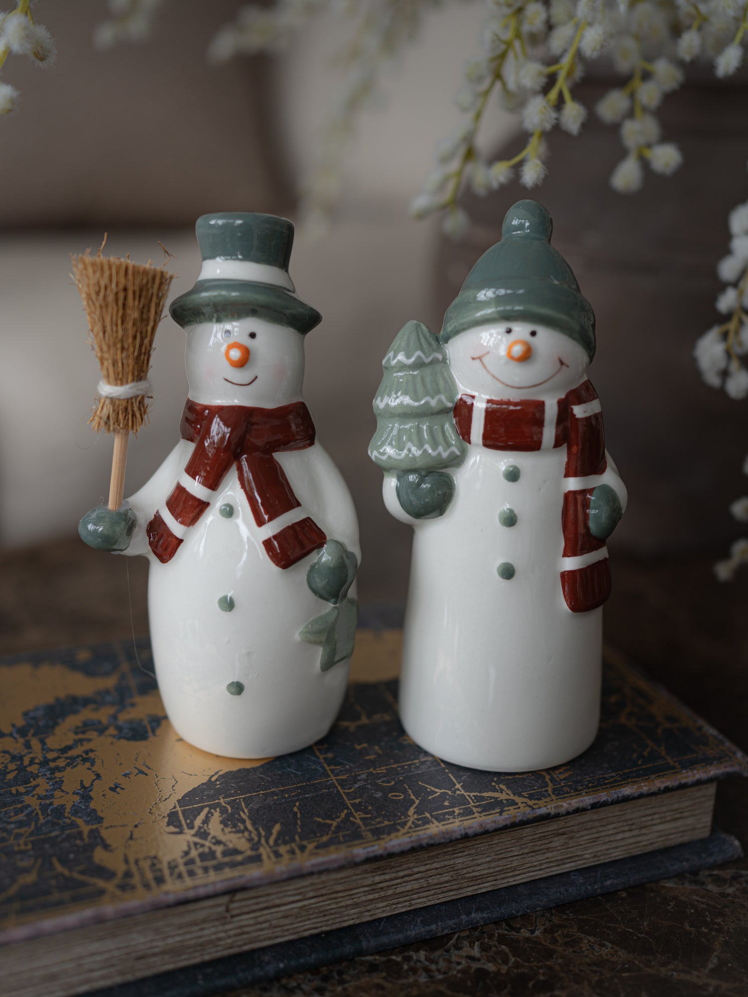 Two white ceramic snowman figurines with green and red holiday accents, displayed on a vintage book with soft-focus flowers in the background.