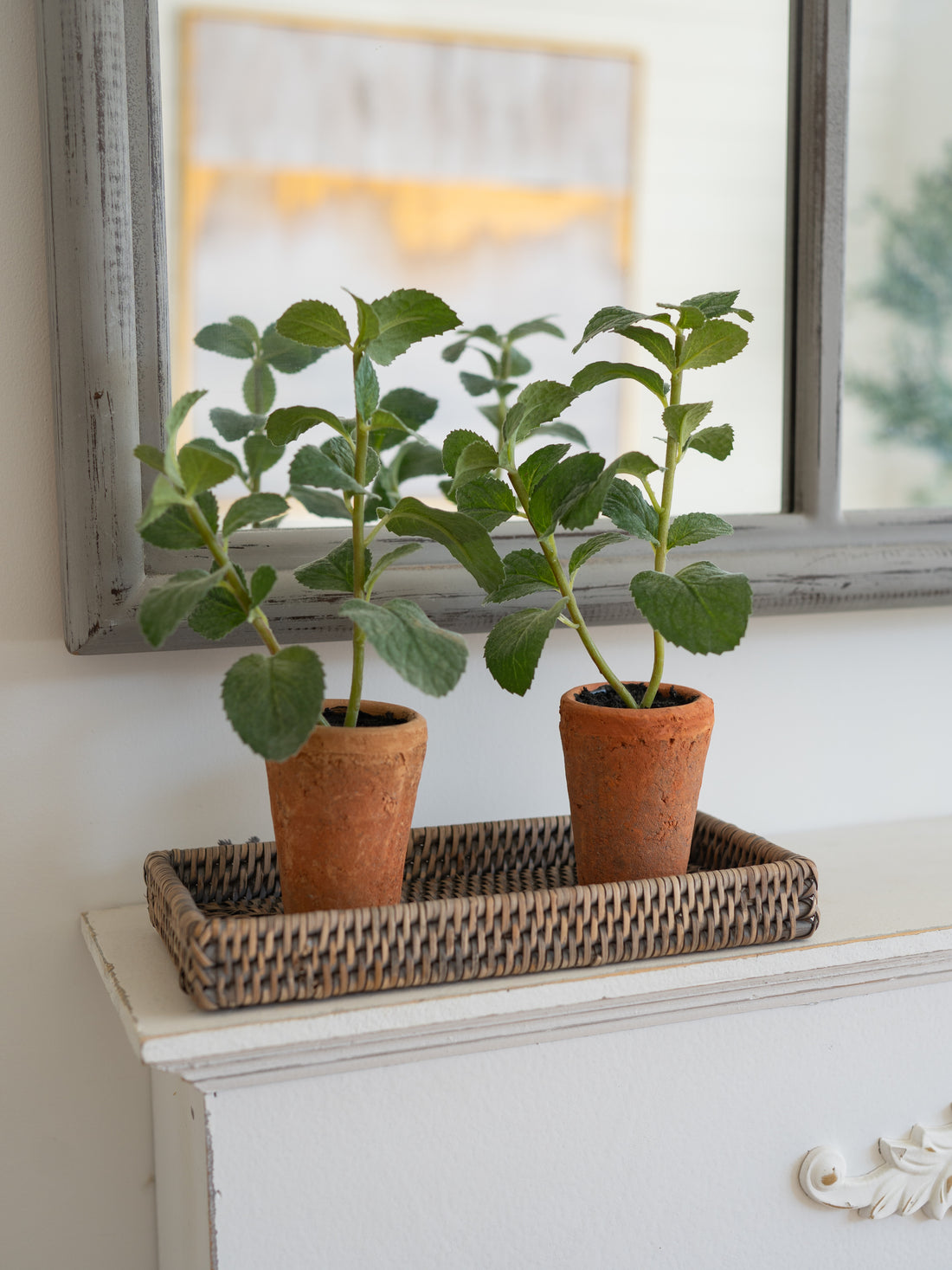 Two potted faux herb plants on a woven tray in front of a framed picture on a white surface.