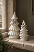 Two white ceramic Christmas tree ornaments with silver bows, displayed on a stone windowsill next to a window and evergreen branches.