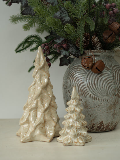 Two ceramic Christmas tree ornaments in cream and off-white glaze, displayed next to a rustic gray patterned vase filled with evergreen branches and pinecones.