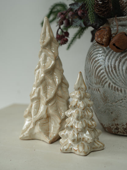 Two small ceramic Christmas tree ornaments in a cream-colored glaze, displayed next to a textured gray ceramic pot with pinecones and berries.