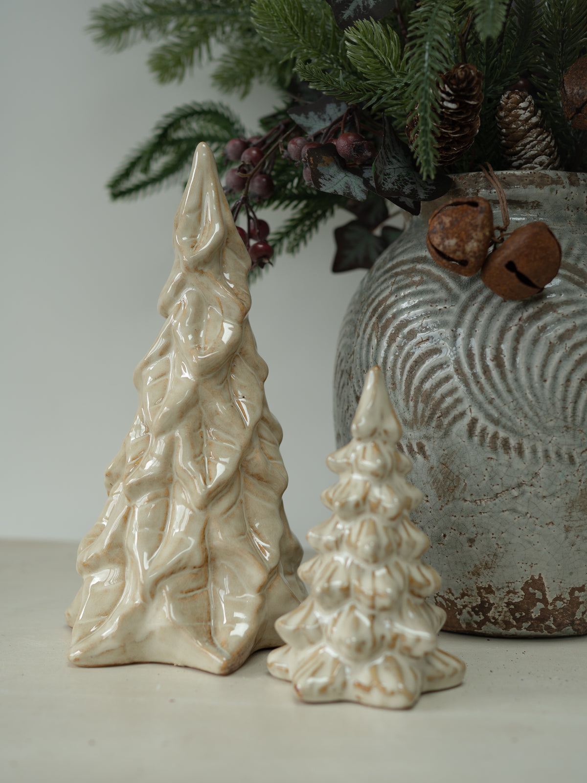 Two small ceramic Christmas tree ornaments in a glossy cream glaze, displayed next to a rustic gray ceramic pot with evergreen branches and bells.