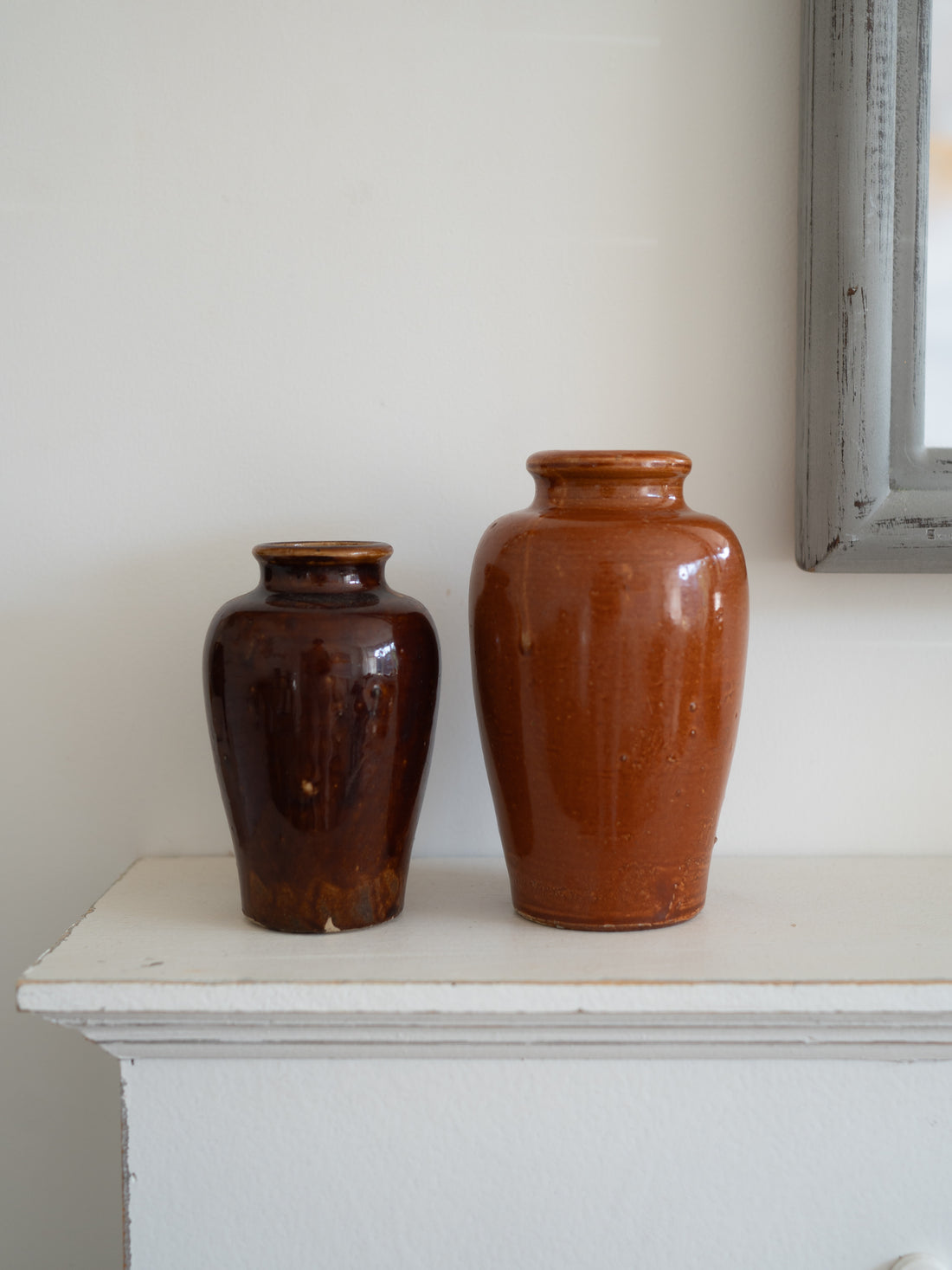 Two glazed ceramic vintage vases, one dark brown and one terracotta orange, displayed on a white mantelpiece against an off-white wall.