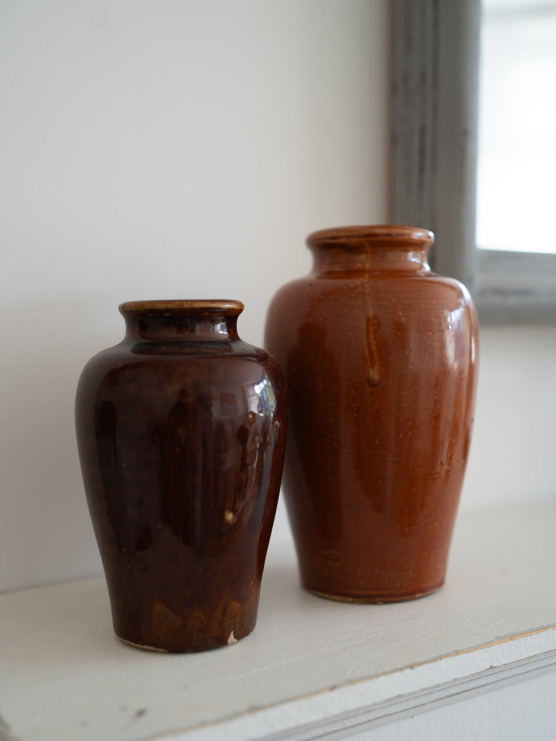 Two small glazed ceramic vintage vases in warm brown tones, one dark reddish-brown and one lighter terracotta brown, standing on a white windowsill.