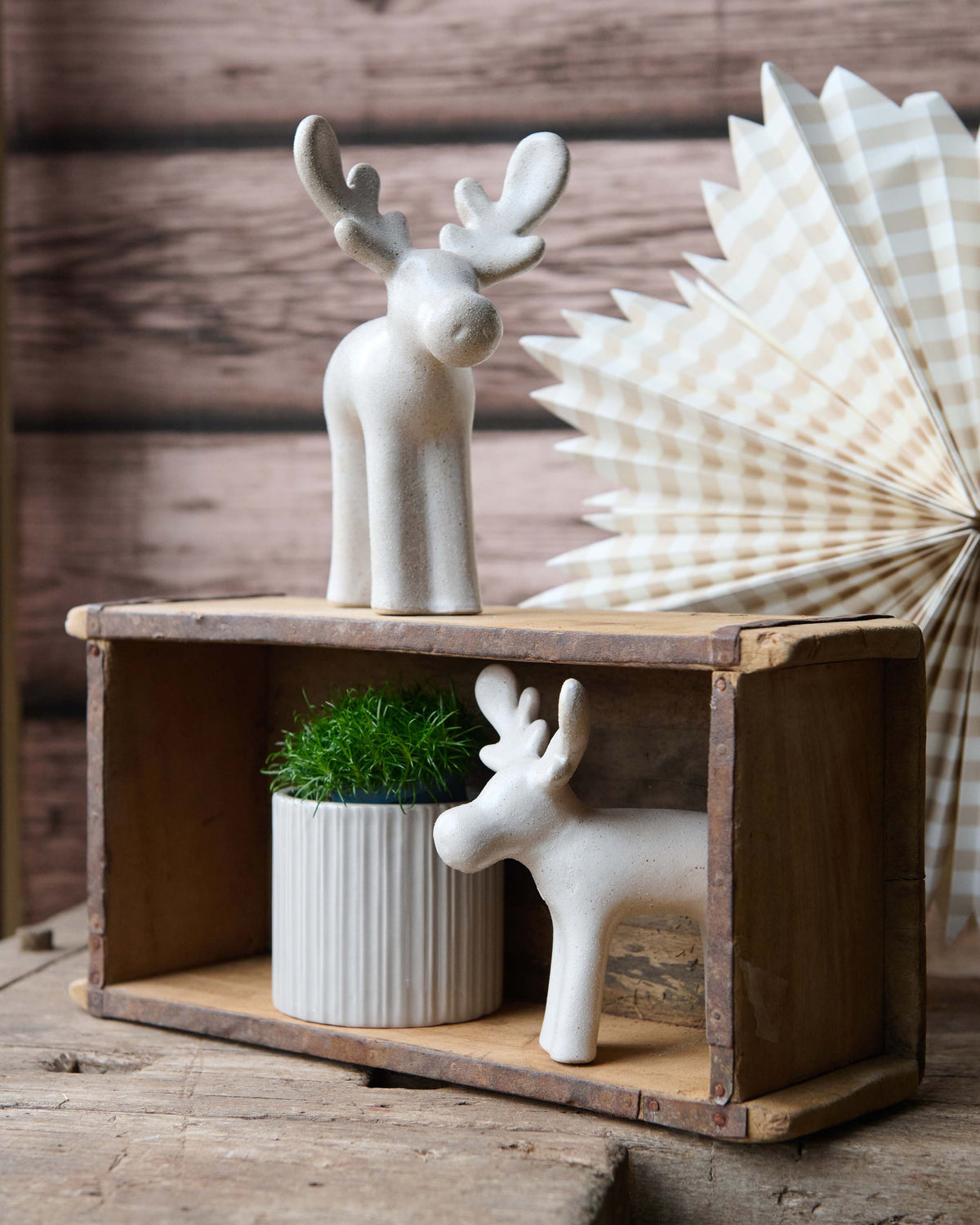 Two white ceramic reindeer figurines displayed on a rustic wooden crate, with one standing on top and one inside the crate next to a small potted green plant. A cream and brown checkered paper fan is visible in the background against a dark wood plank wall.