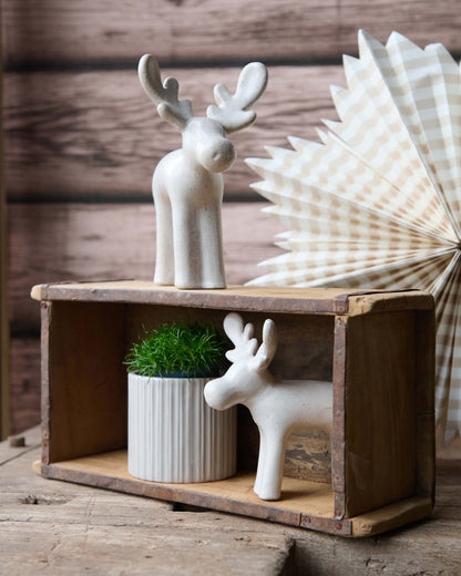 Two white ceramic reindeer figurines displayed on a rustic wooden crate, with one standing on top and one inside the crate next to a small potted green plant. A cream and brown checkered paper fan is visible in the background against a dark wood plank wall.