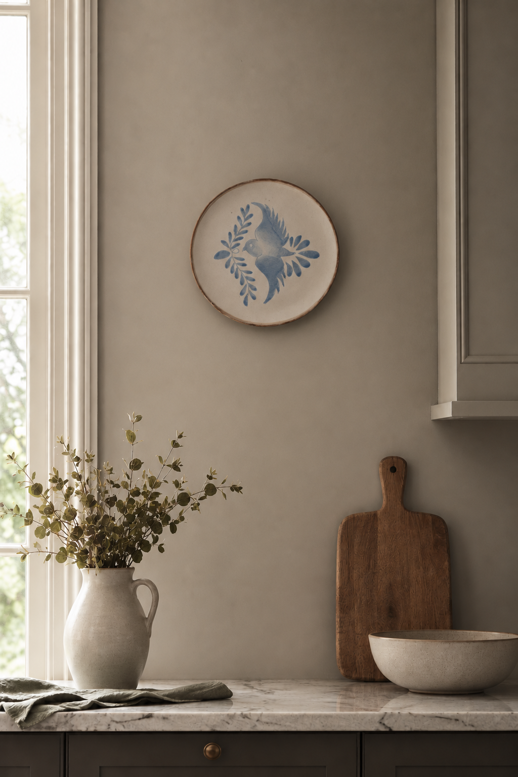 Decorative plate with floral  and songbird design on a wall above a kitchen counter with a vase, cutting board, and bowl.