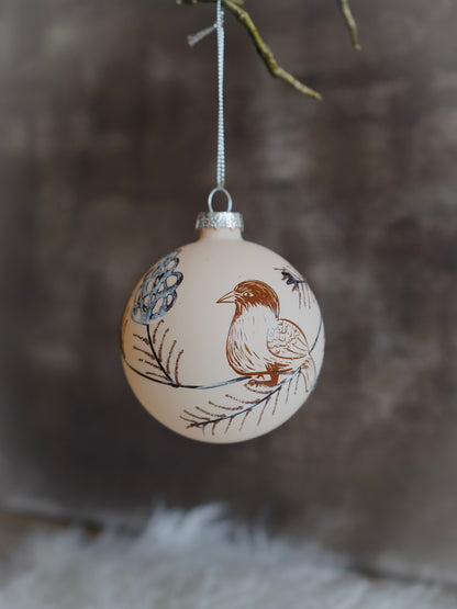 Decorative Christmas ball ornament with bird and floral design hanging against a blurred natural background.