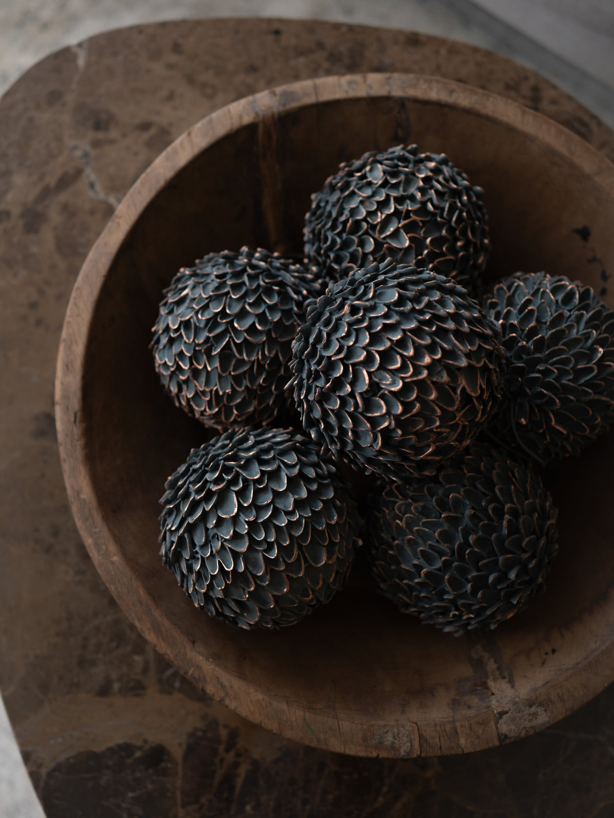 Five dark, textured decorative spheres with a pine cone pattern, arranged inside a rustic wooden bowl on a stone surface.