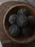 Five dark, textured decorative spheres with a pine cone pattern, arranged inside a rustic wooden bowl on a stone surface.