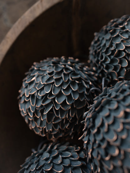 Close-up of a decorative sculptural object with a dark brown and charcoal, pine cone-like textured surface, resting inside a dark wooden bowl.