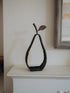 A dark metal decorative sculpture shaped like a pear with a leafy stem, displayed on a white mantelpiece against a warm off-white wall.