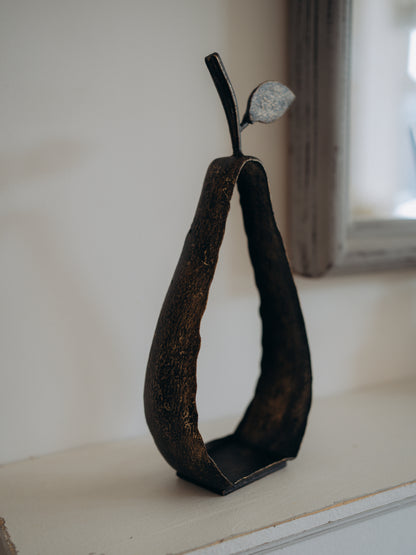 A dark metal decorative sculpture shaped like a pear with a textured, rustic finish and a small leaf on top, displayed on a light-colored windowsill.