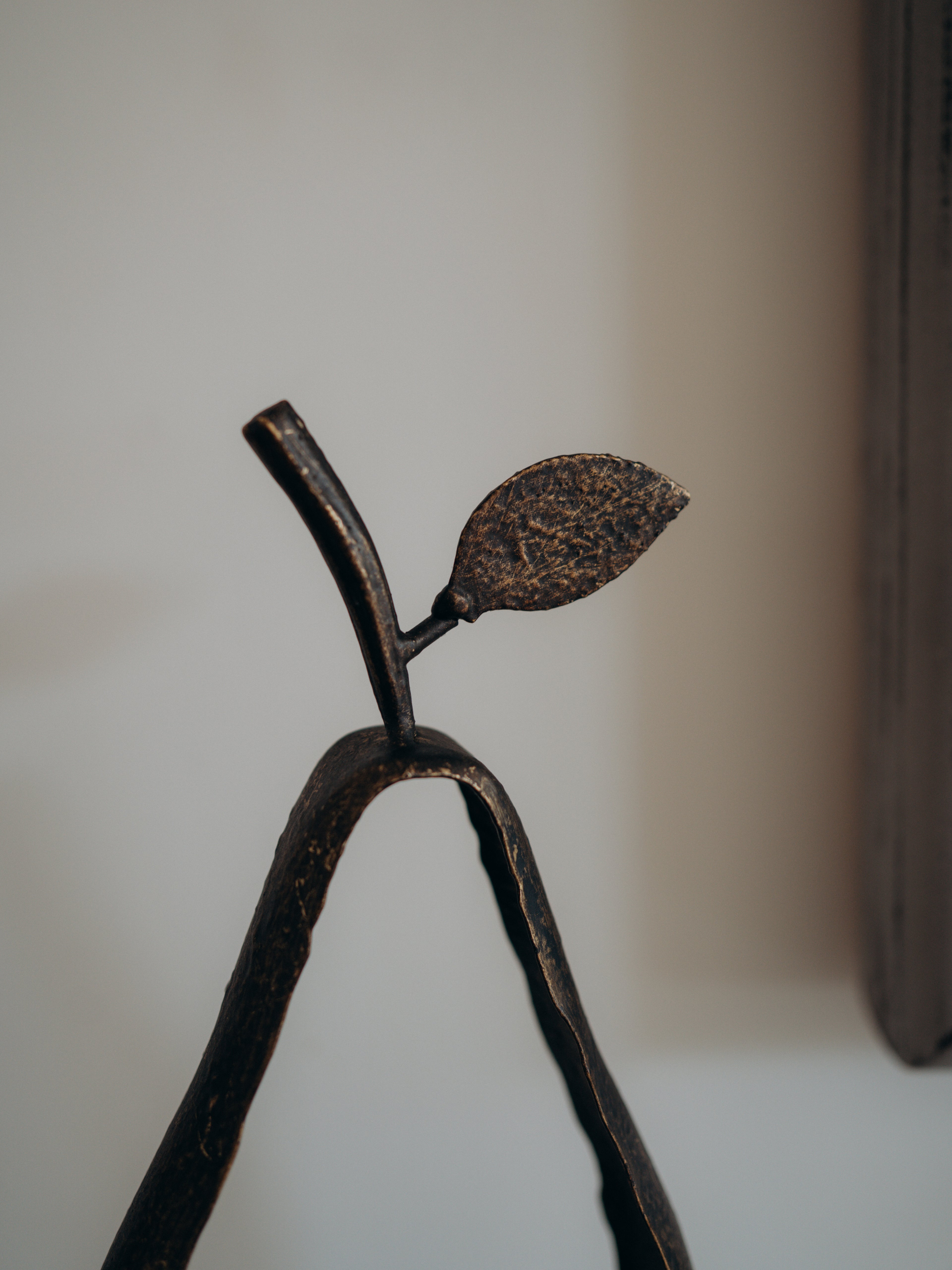 A close-up of a dark metal decorative sculpture shaped like a stylized pear with a leafy stem, photographed indoors against a soft beige wall.
