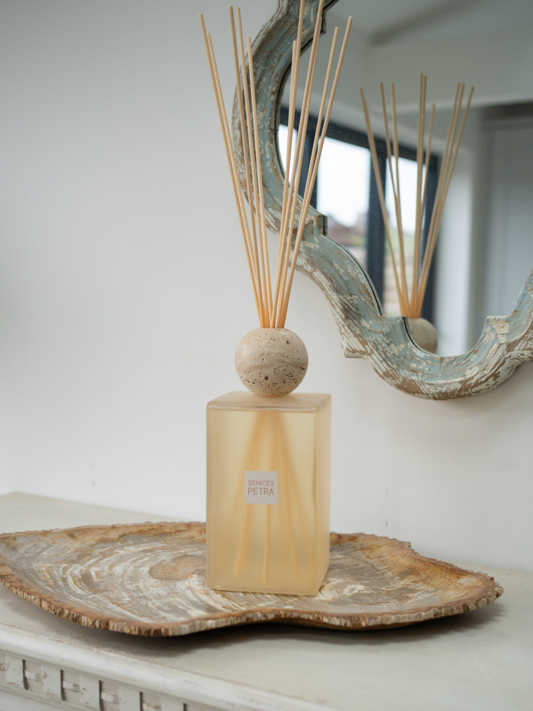 A extra large statement petra reed diffuser bottle with a frosted beige-tinted glass body, a round beige stone top, and natural reed sticks, displayed on a rustic wood slice tray against a white wall with a decorative mirror.