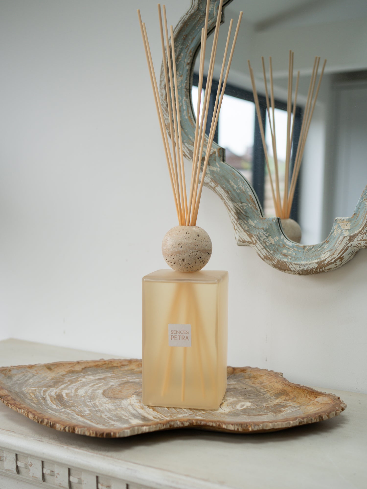 A extra large statement petra reed diffuser bottle with a frosted beige-tinted glass body, a round beige stone top, and natural reed sticks, displayed on a rustic wood slice tray against a white wall with a decorative mirror.