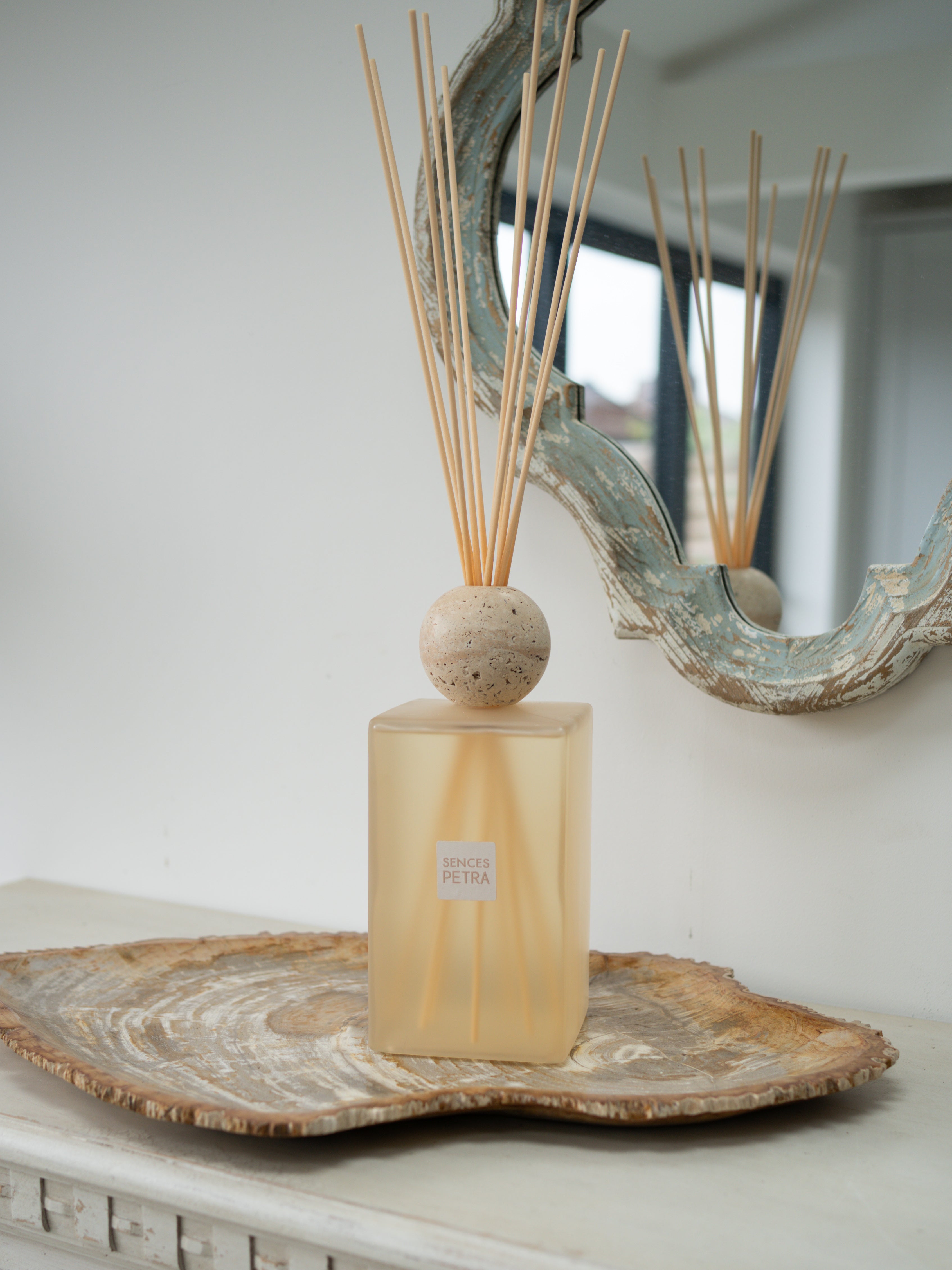 A extra large statement petra reed diffuser bottle with a frosted beige-tinted glass body, a round beige stone top, and natural reed sticks, displayed on a rustic wood slice tray against a white wall with a decorative mirror.