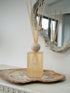 A extra large statement petra reed diffuser bottle with a frosted beige-tinted glass body, a round beige stone top, and natural reed sticks, displayed on a rustic wood slice tray against a white wall with a decorative mirror.
