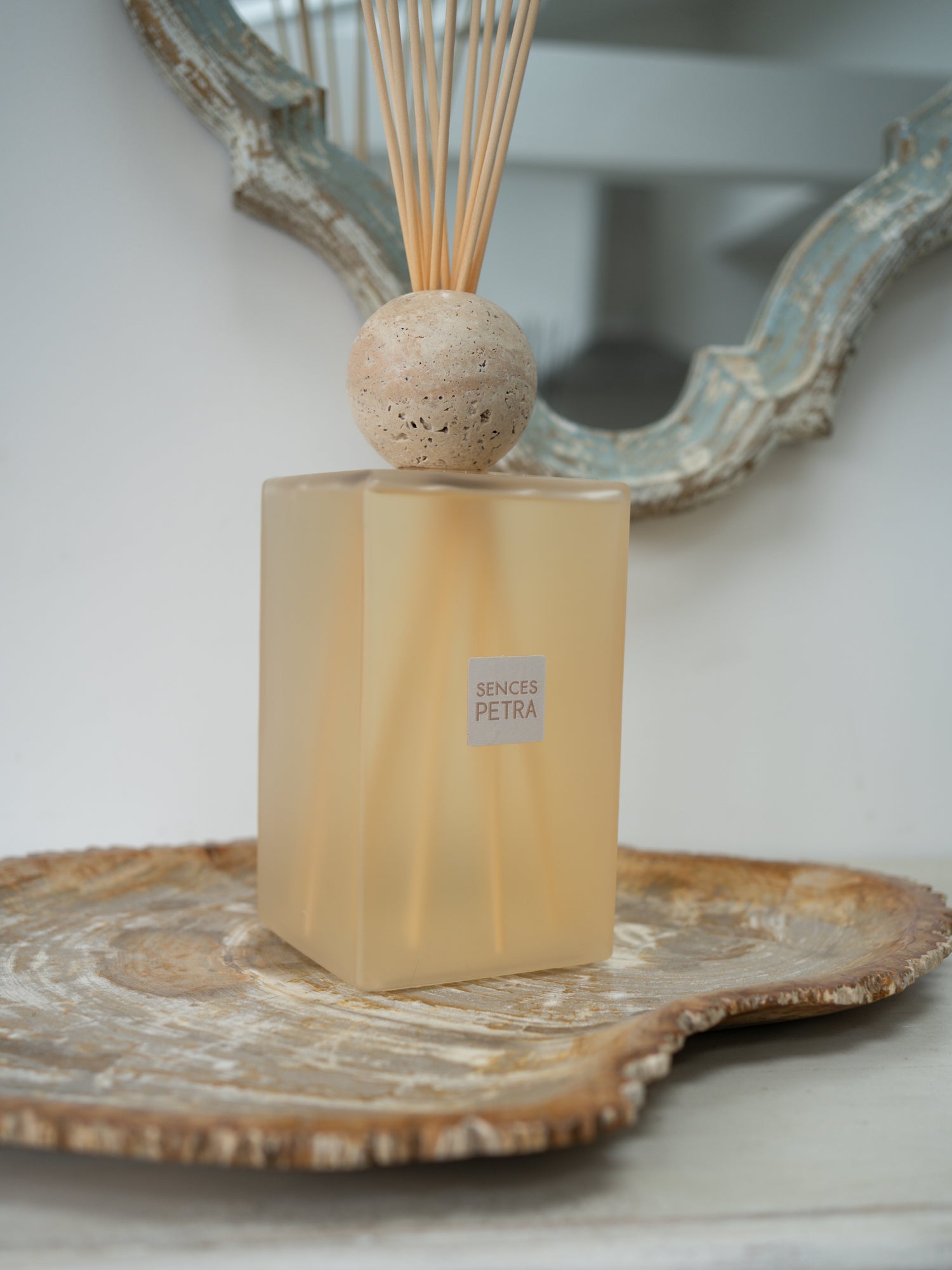 A reed diffuser bottle with a frosted pale blush body, a round natural travertine stone top, and reeds, displayed on a decorative tray with a distressed mirror in the background.