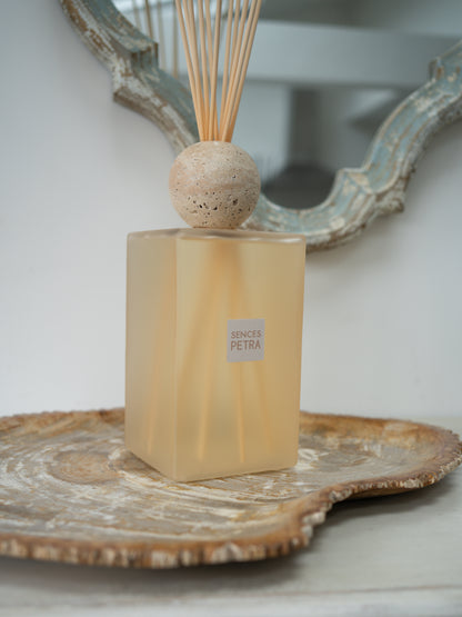 A reed diffuser bottle with a frosted pale blush body, a round natural travertine stone top, and reeds, displayed on a decorative tray with a distressed mirror in the background.